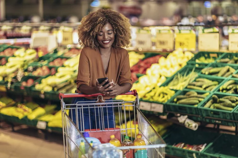 mulher negra caminha sorridente em mercado com celular em mãos